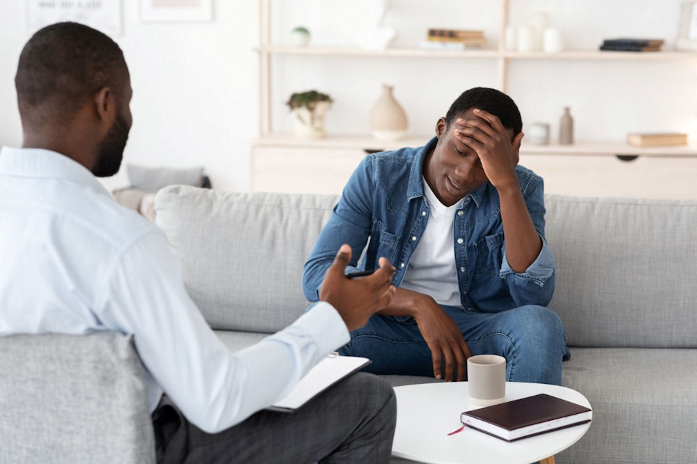Man speaking with a therapist while holding his head, representing emotional strain and support needs tied to perfectionism and mental health.
