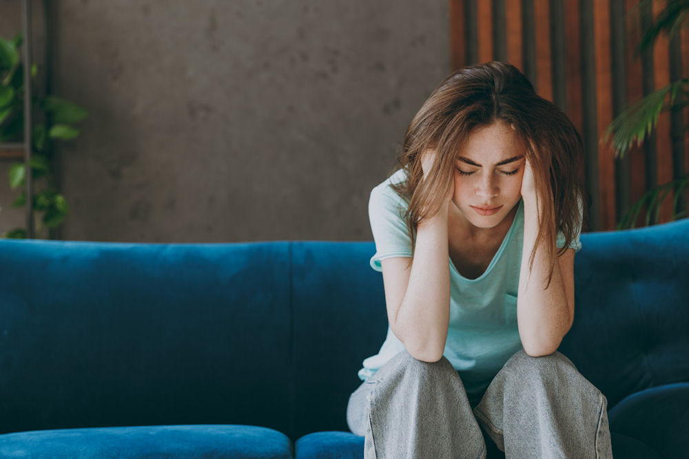Young woman sitting on a couch looking stressed and contemplative, illustrating challenges for women in recovery.